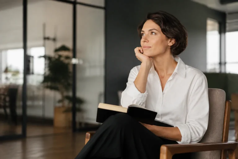 A confident woman in a modern office setting, engaging in conversation with relaxed body language and warm natural light illuminating the scene.