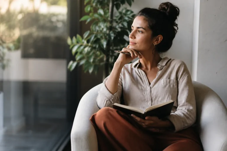 A thoughtful woman sits at a modern desk with a notebook, looking contemplatively out a large window as warm light illuminates her face.